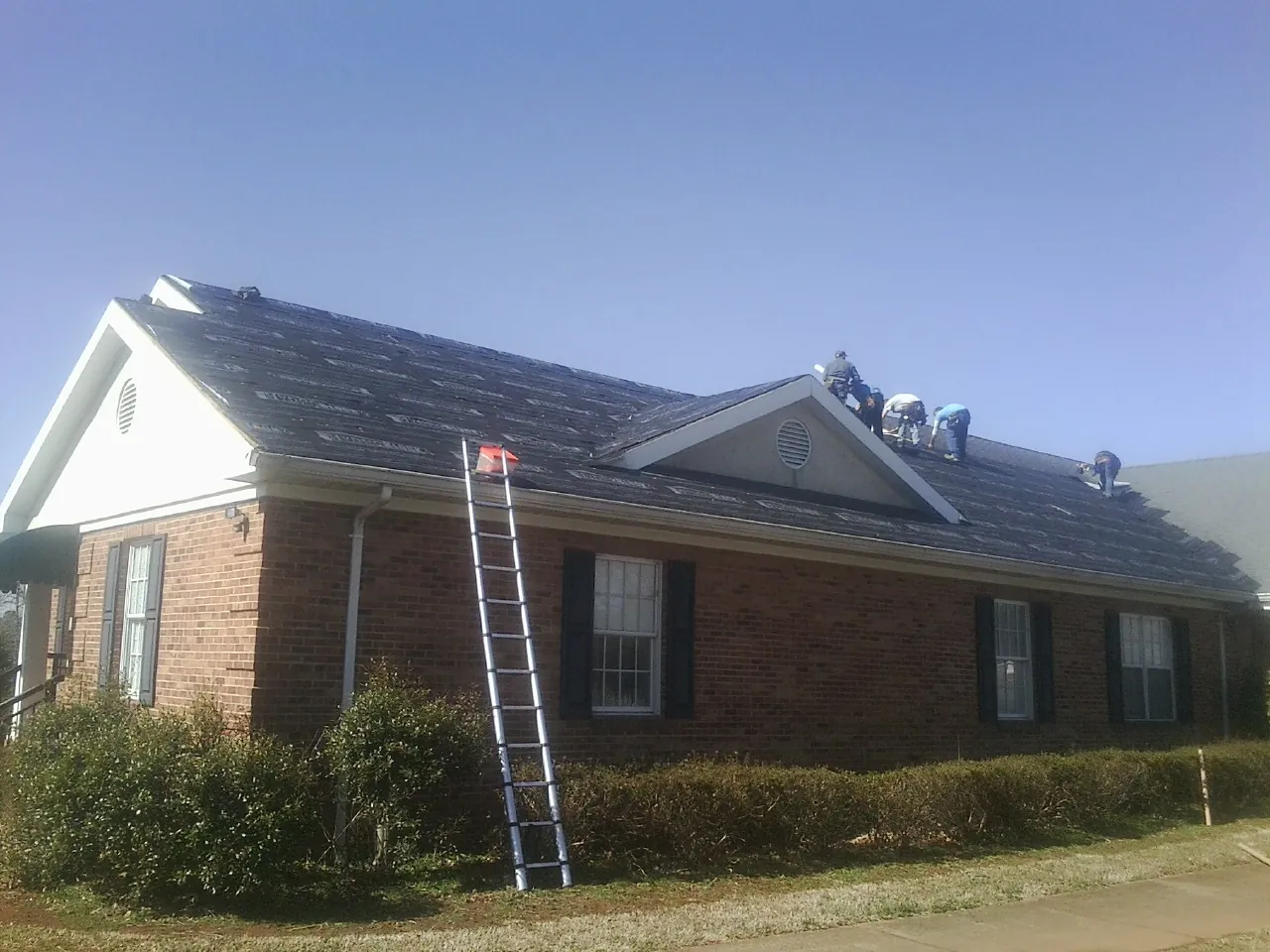 Crew tearing shingles off a brick ranch roof with a ladder set — Metal Roof Repair work by our crew in Dell