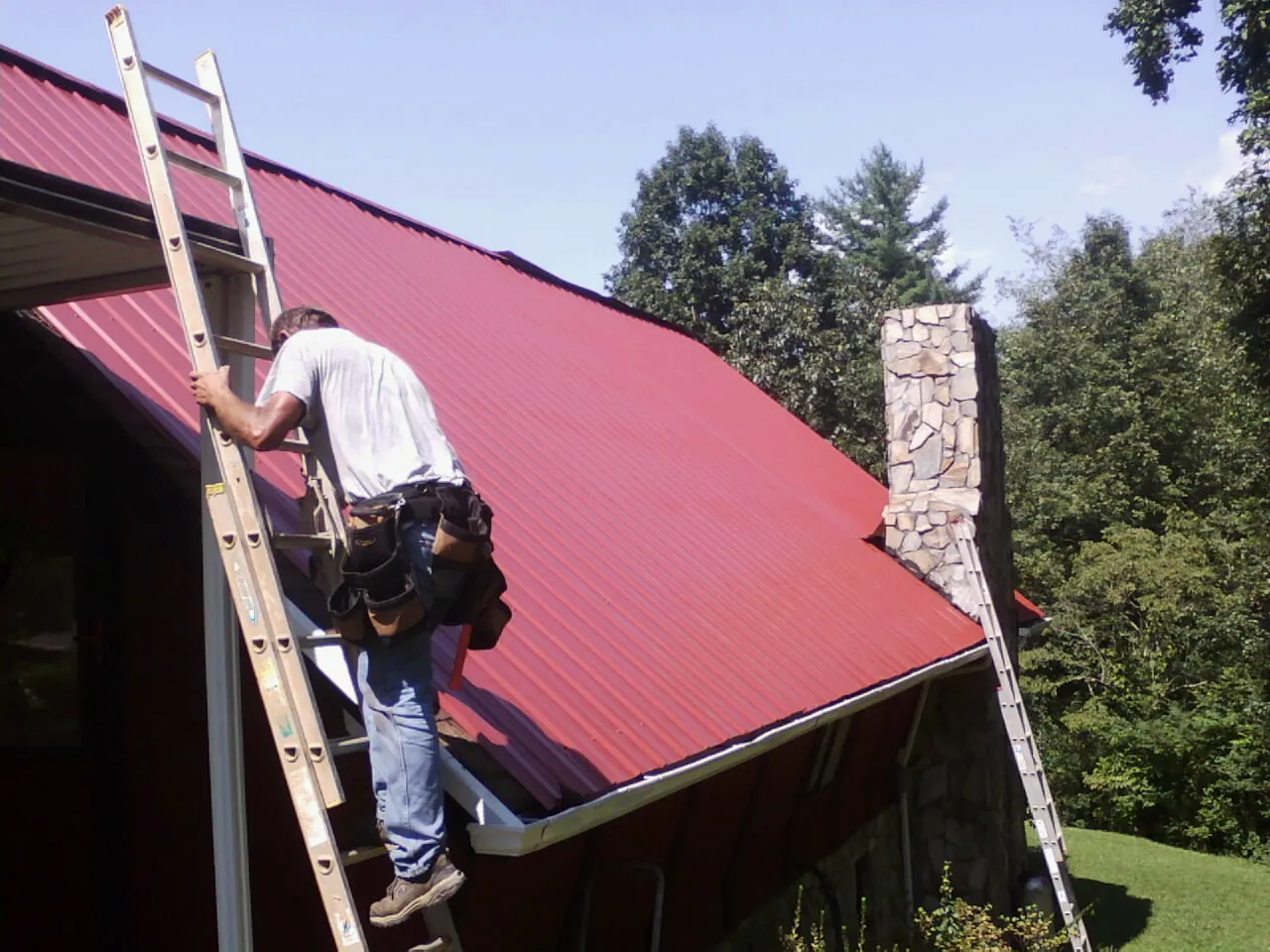 Roofer on a ladder beside a red metal roof and stone chimney — Soffit & Fascia Repair by Roofing Brothers in Dell